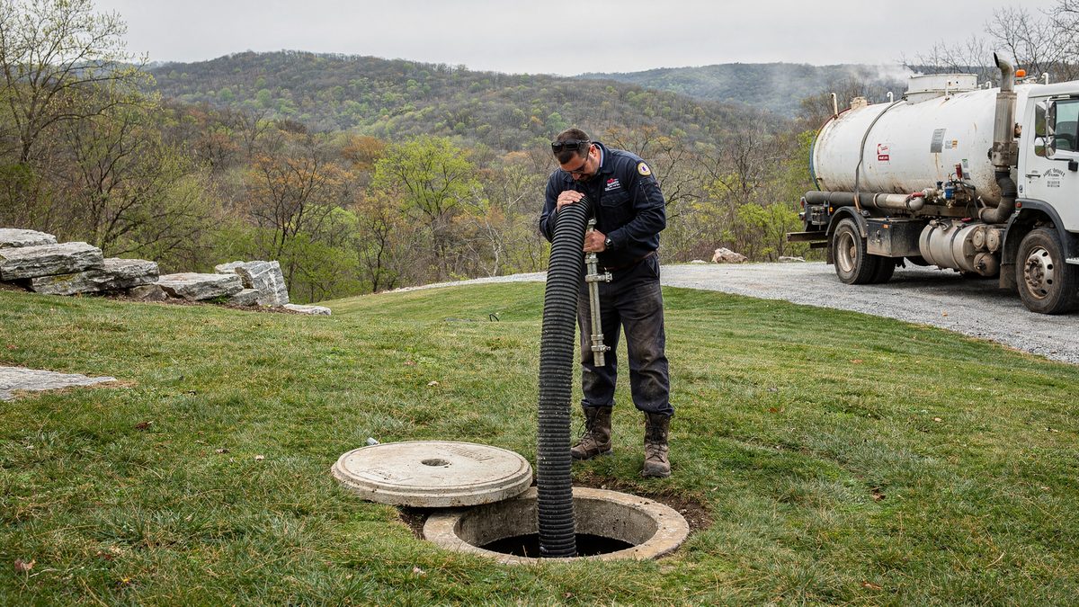 Septic technician steadying a corrugated vacuum hose into an open concrete septic tank in a rural Greene County, Missouri yard, the lid set aside and the vacuum truck staged on the gravel drive behind